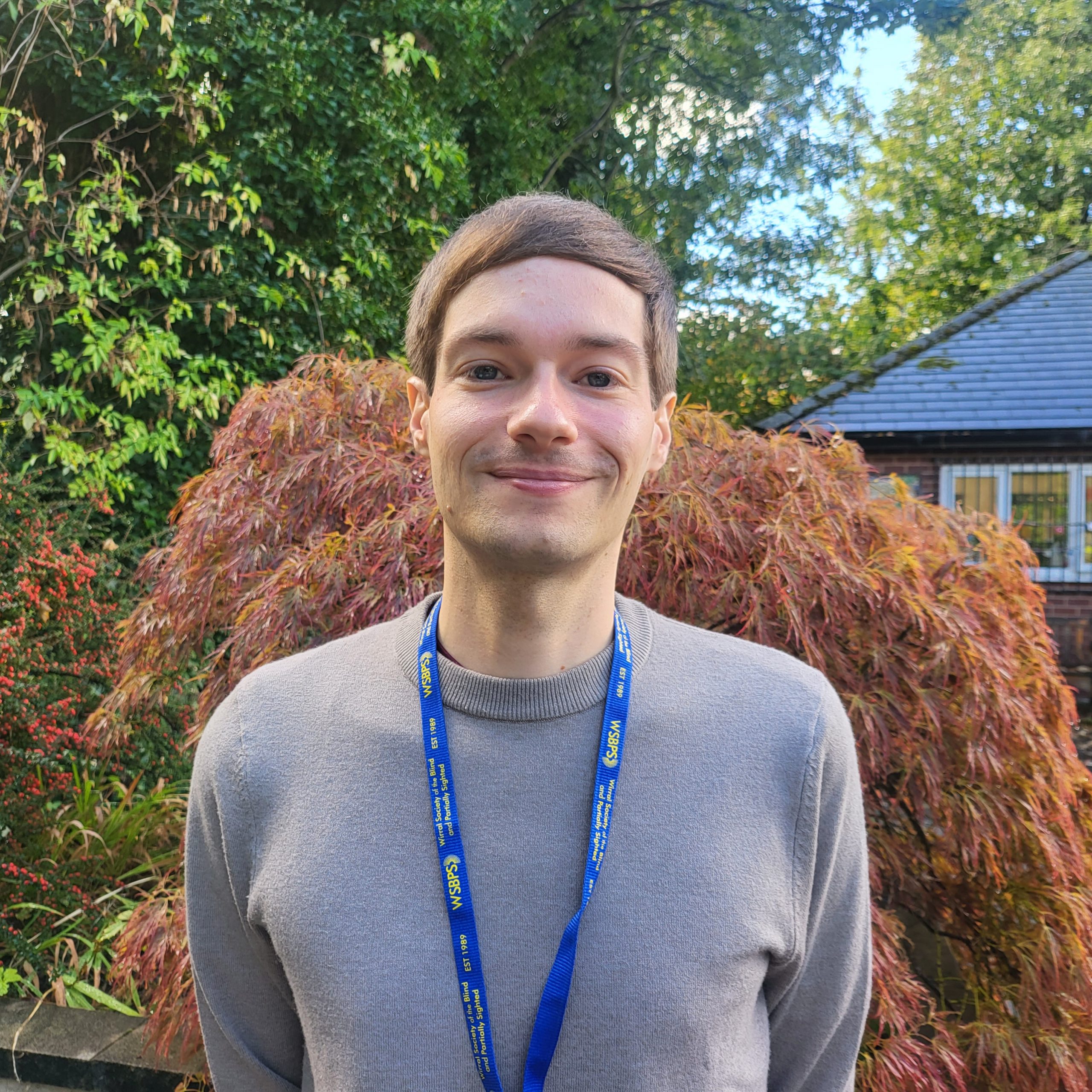 A man with neat brown hair is standing in the sensory garden at Ashville Lodge, he is smiling and wearing a grey jumper.