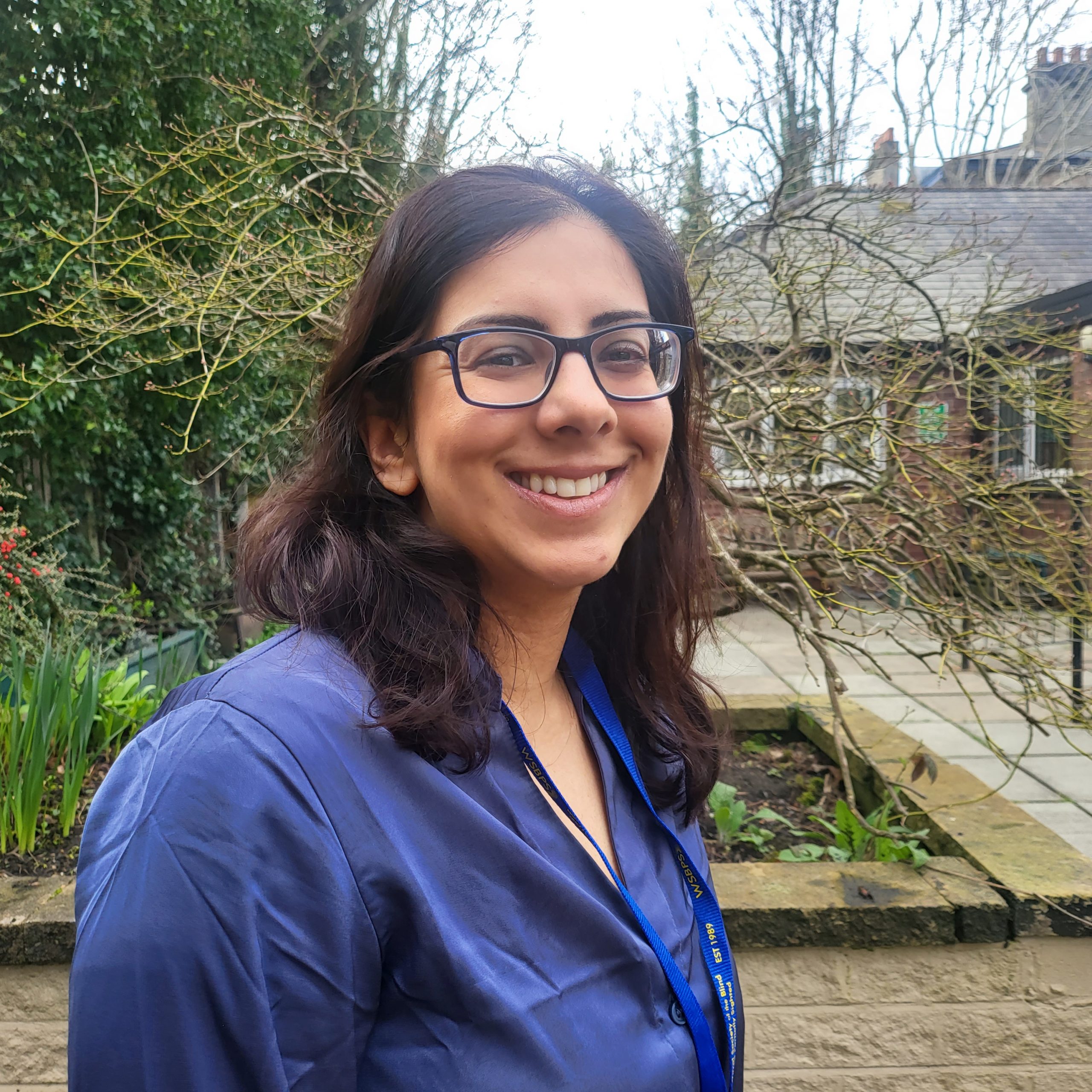 20260317_110344 A woman with shoulder length dark brown hair is standing in the sensory garden at Ashville Lodge, she is smiling and wearing black-rimmed glasses and a dark blue shirt.
