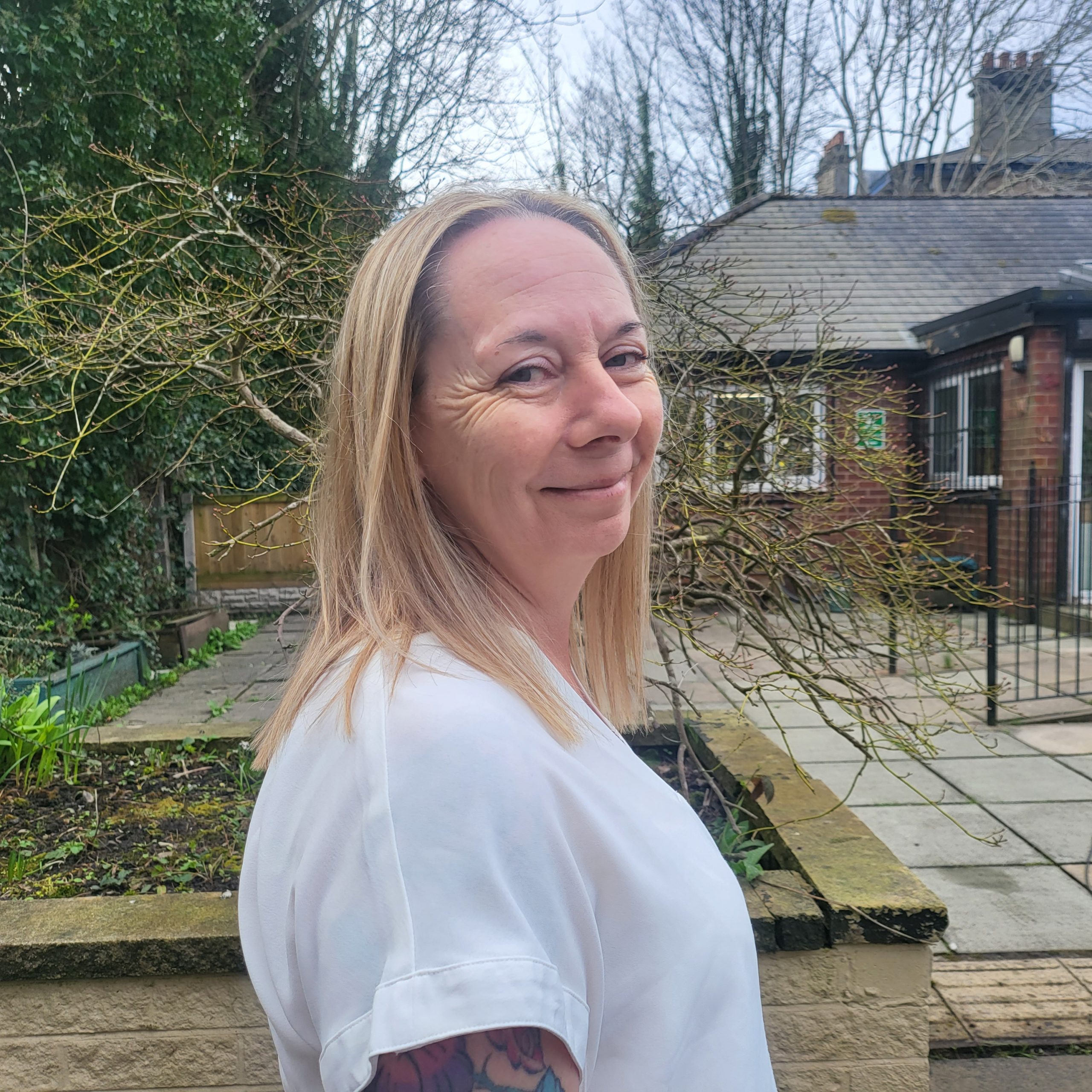 20260317_110509 A woman with shoulder length blonde hair is standing in the sensory garden at Ashville Lodge, she is smiling and wearing a white top with short sleeves.