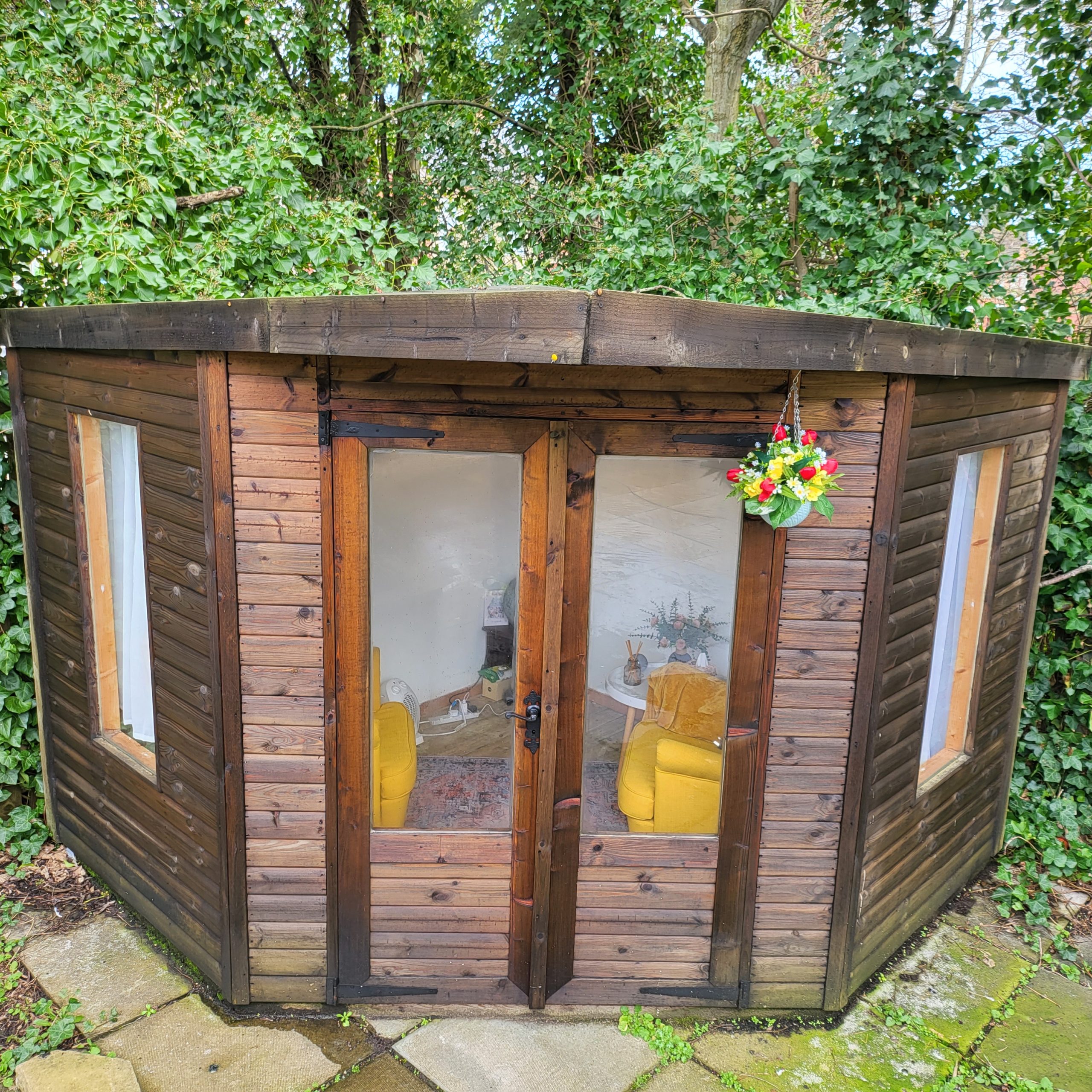 The summerhouse where our counsellor holds counselling sessions. The summerhouse is made of dark stained wood, through the windows can be seen two bright yellow comfy armchairs.