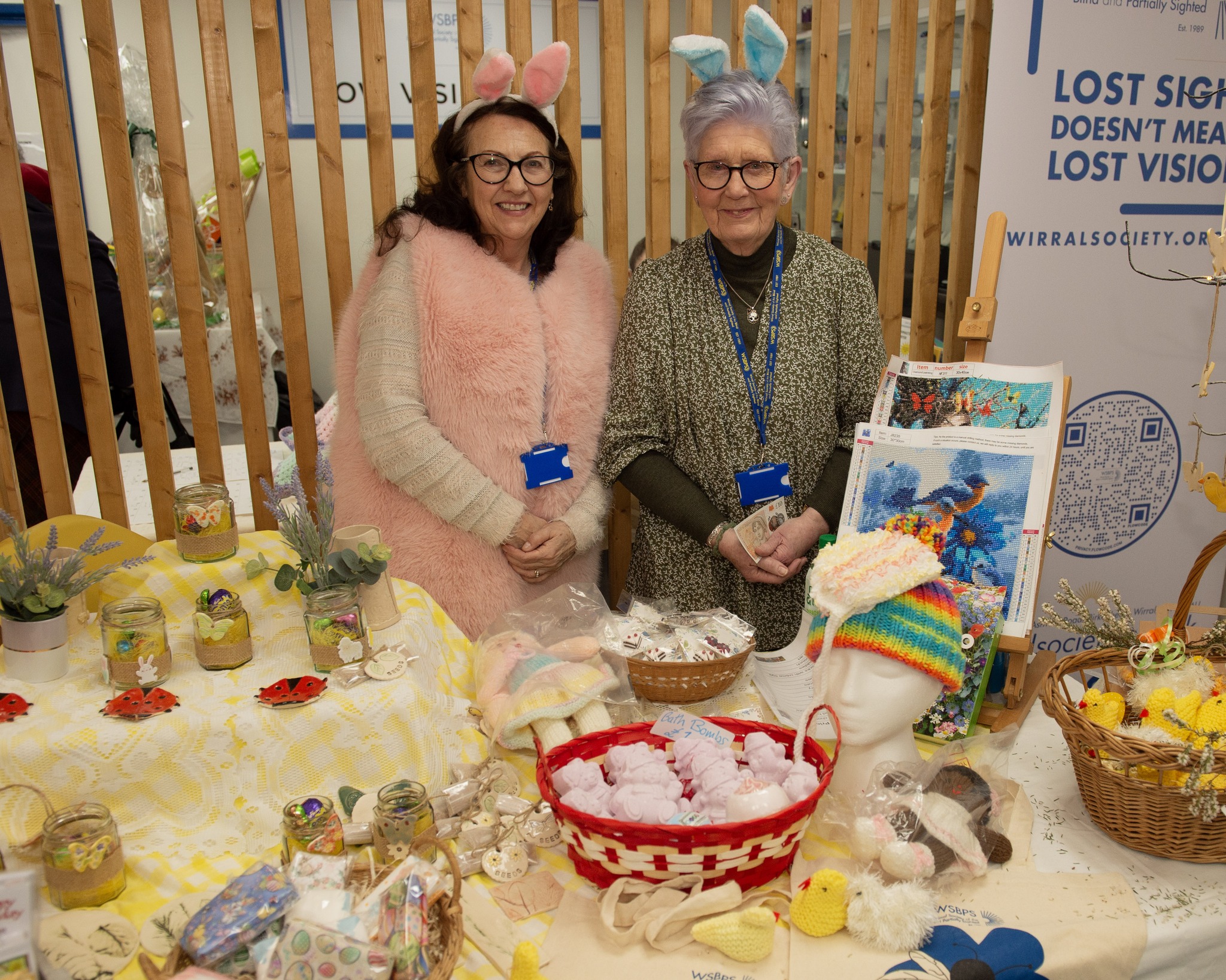 484398482_1055750756586916_7418891237056130710_n Two female volunteers running a craft stall at an Easter fair, they are wearing bunny ears and one of the volunteers is wearing a long, fluffy pink top.