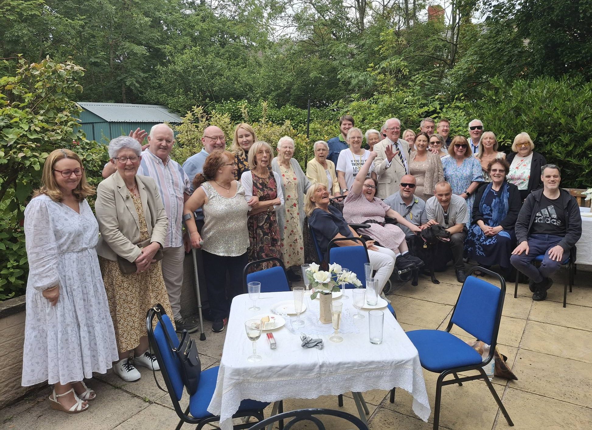 485727540_1061151632713495_16045292774751263_n A group of volunteers dressed in summer clothes are at a social gathering in the sensory garden at Ashville Lodge, they are smiling whilst waving at the camera.