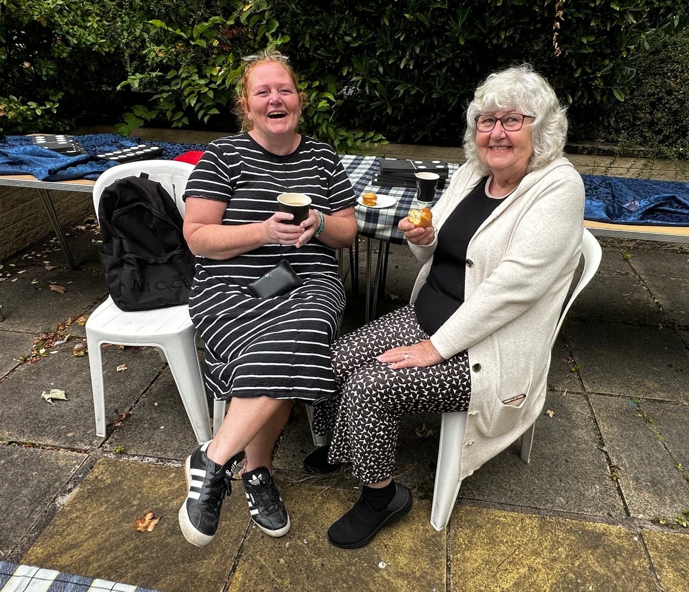 550314990_1206812834814040_3436173290908113269_n Two women are sitting on plastic garden chairs on the patio in the sensory garden at Ashville Lodge, they are holding drinks and food whilst smiling at the camera.