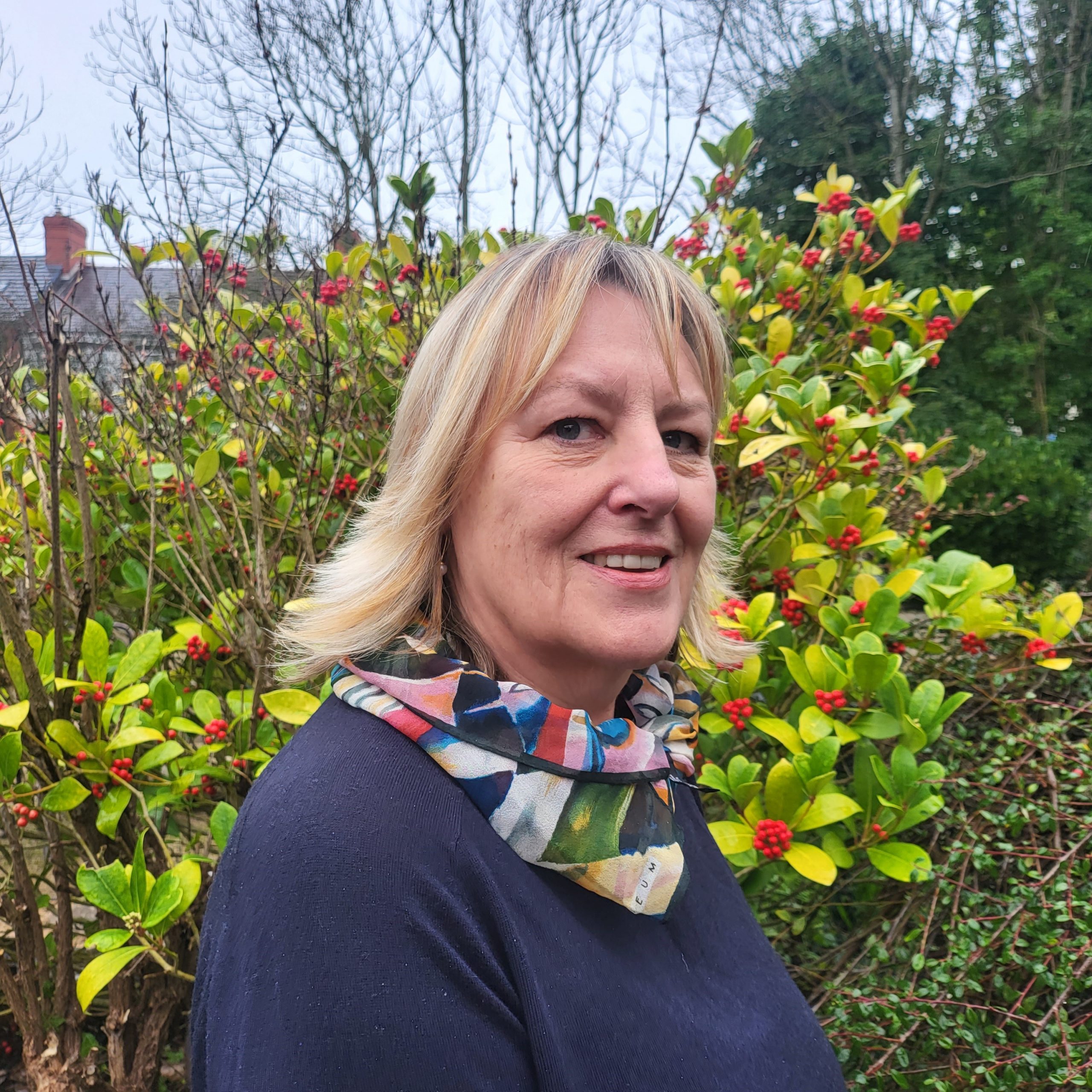 Adele Law – Trustee – Director A woman with shoulder length blonde hair is standing in the sensory garden at Ashville Lodge, she is smiling and wearing a small, brightly coloured scarf over a navy top.