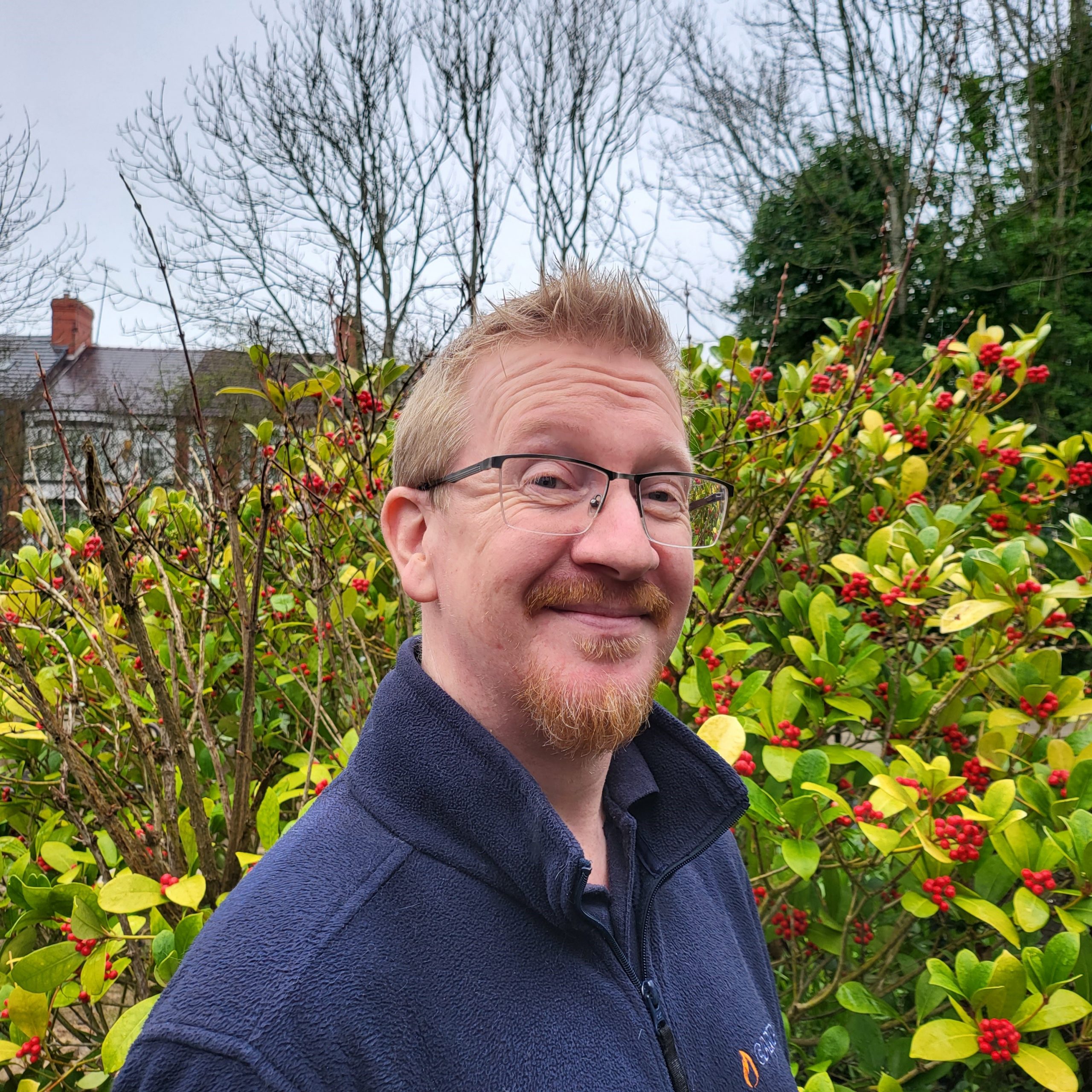 Chris Kelly – Trustee – Director A cheerful man with short ginger hair and a goatee beard is standing in the sensory garden at Ashville Lodge, he is wearing glasses and a dark blue fleece jacket.