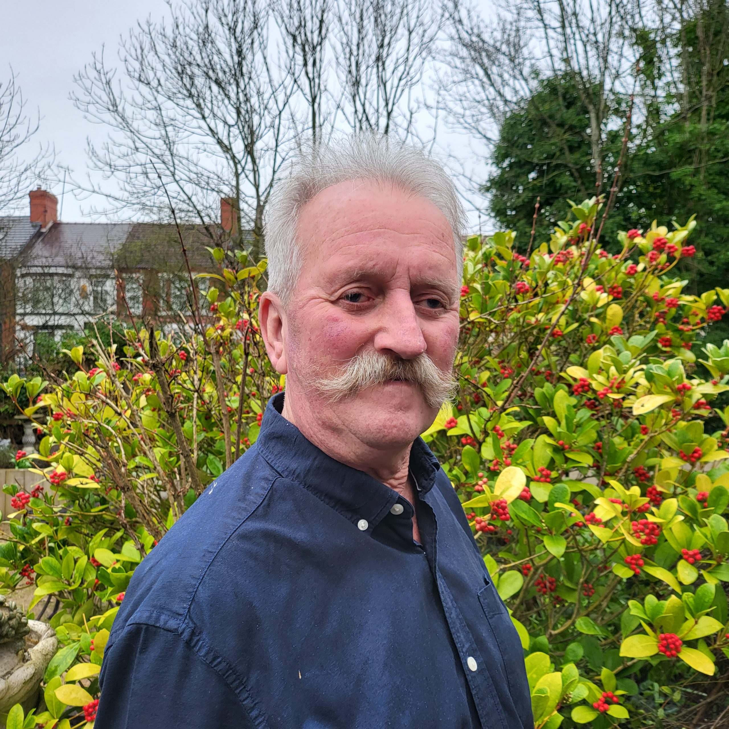 Gary – Trustee – Director A man with short grey hair and a handlebar moustache is standing in the sensory garden at Ashville Lodge, he is smiling and wearing a navy shirt with white buttons.