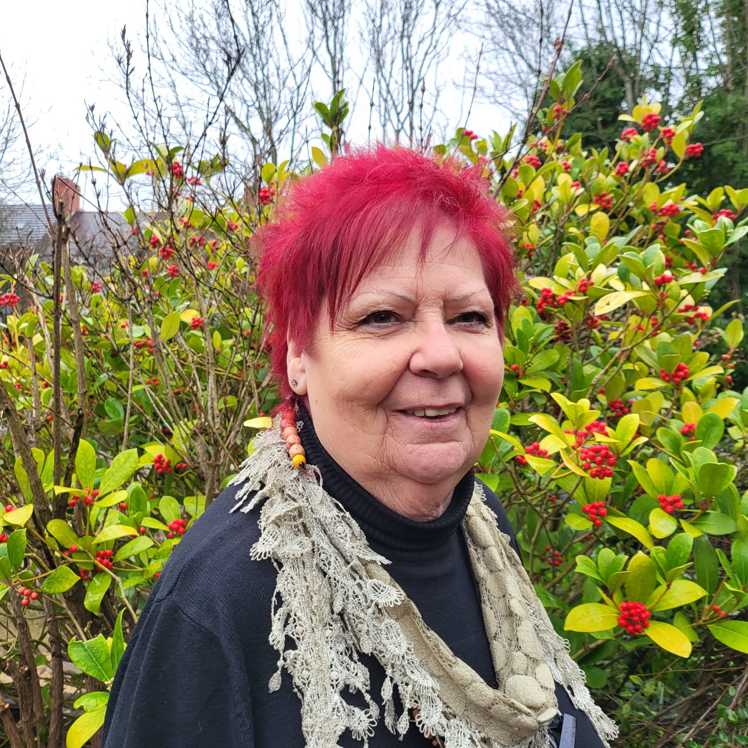 Lynn – Trustee – Director A smiling woman with shortish bright red hair is standing in the sensory garden at Ashville Lodge, she is wearing brightly coloured dangly earrings and a delicate, white lace scarf.