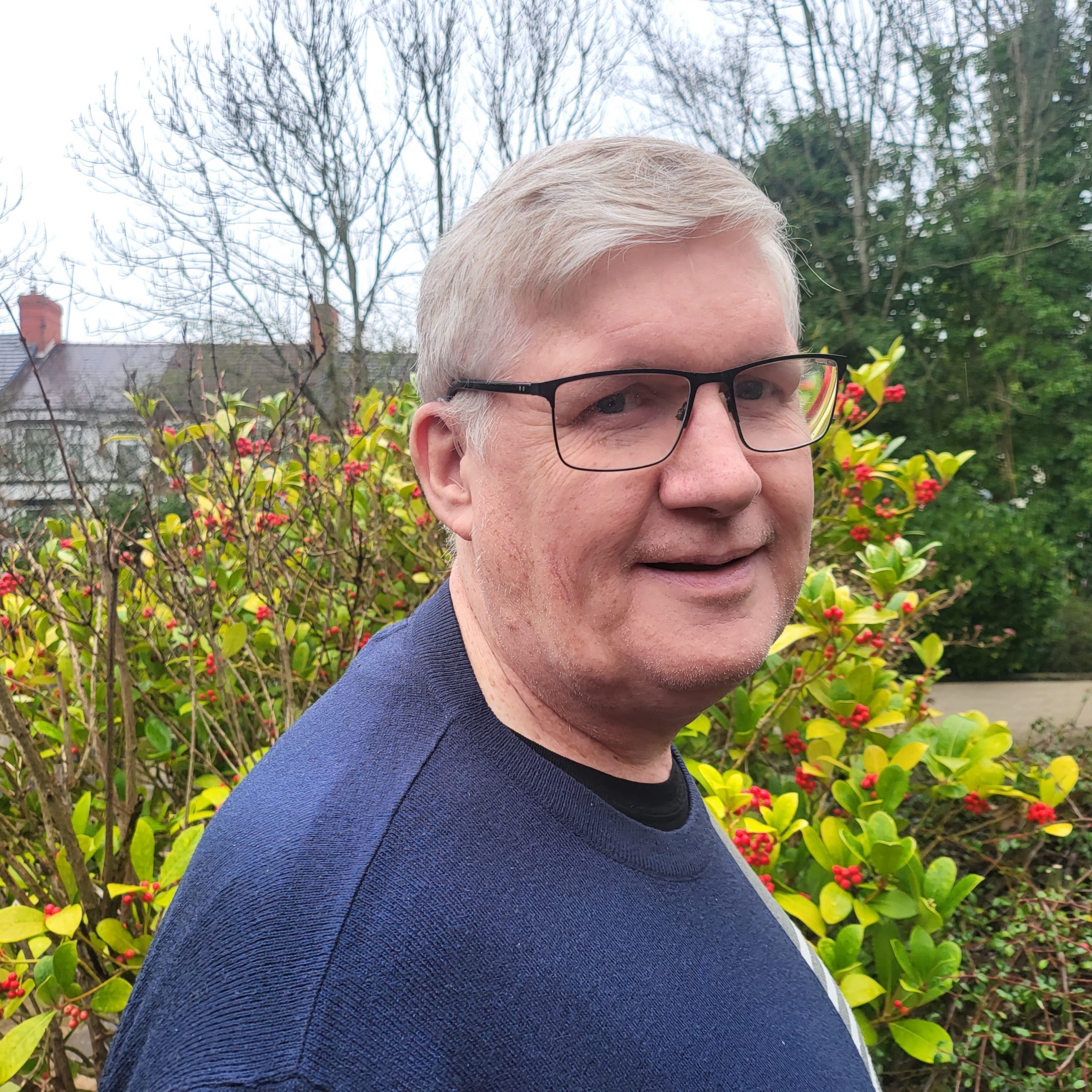 TBC Trustee – Director 1 A smiling man with shortish grey hair is standing in the sensory garden at Ashville Lodge, he is wearing black-rimmed glasses and a dark blue jumper.
