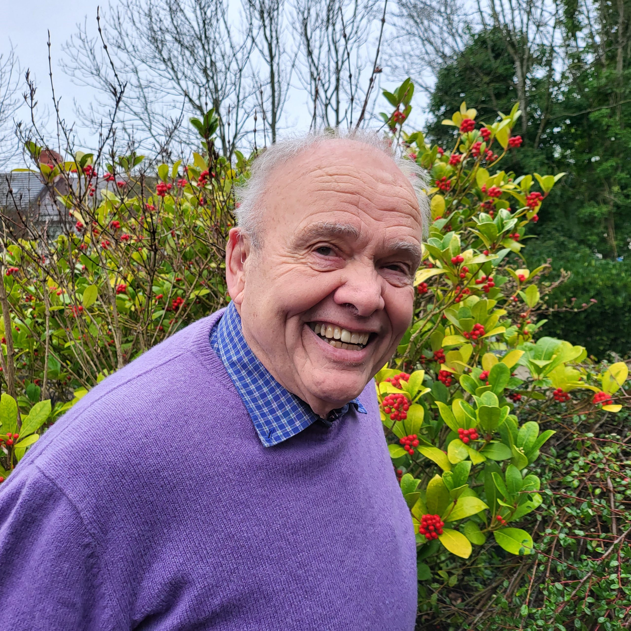 TBC Trustee – Director A man with short grey hair is standing in the sensory garden at Ashville Lodge, he has a cheeky smile and is wearing a purple jumper.