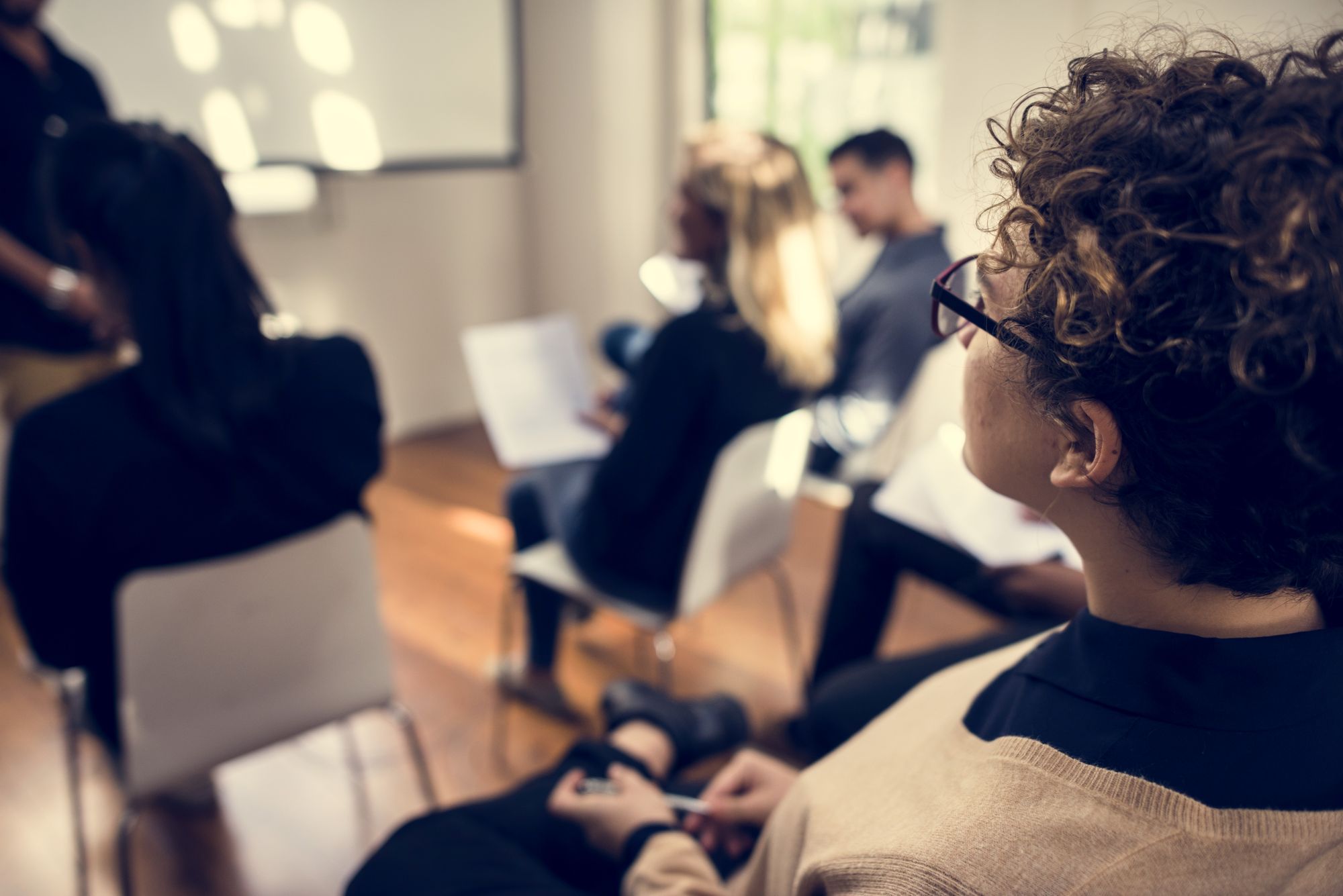 business-people-in-a-meeting-2026-01-07-23-58-04-utc A small group of people are sitting on chairs in rows in an office, they are listening attentively to a presentation being given by a speaker at the front of the room.