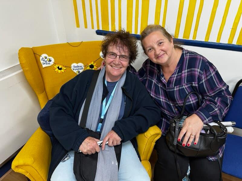 Two women are sitting close together in Ashville Lodge, they are leaning towards each other and smiling at the camera. The woman on the left is sitting on a bright yellow armchair, she has cheerfully windswept hair and is wearing clothes in various shades of blue. The woman on the right is sitting on a low chair, she has neatly tied back hair and is wearing a navy shirt with a blue and grey check pattern.